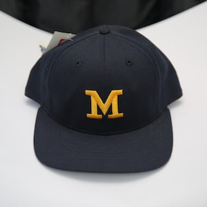 May include: Navy blue baseball cap with a flat brim. The cap features a gold "M" embroidered on the front. The hat has a structured crown and a tag attached to the side. A classic style.