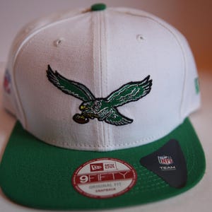 Op de afbeelding: Witte en groene Philadelphia Eagles 9FIFTY snapback pet. De pet heeft het logo van het team, een adelaar met uitgespreide vleugels, en het NFL-logo. De klep is groen en de kroon is wit.