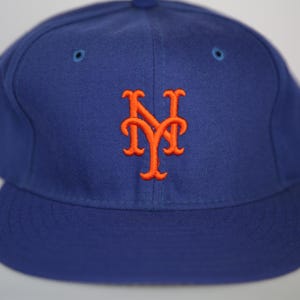 Może przedstawiać: Królewska niebieska czapka bejsbolowa z płaskim daszkiem. Czapka posiada pomarańczowe haftowane logo "NY" z przodu. Czapka ma strukturalną koronę i dwa małe niebieskie oczka na górze.
