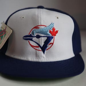 May include: A baseball cap with a white front panel and a navy blue brim and back. The cap features an embroidered logo of a blue bird, a red maple leaf, and a baseball. A tag is attached to the side.