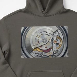 May include: A gray hoodie featuring a close-up image of a watch mechanism. The intricate details of the watch's internal components, including gears and springs, are visible. The hoodie has a kangaroo pocket and a hood.