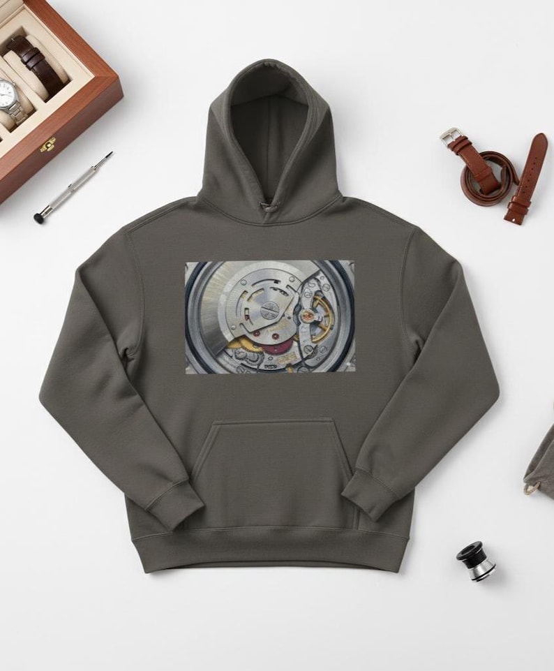 May include: A gray hoodie featuring a close-up image of a watch mechanism. The intricate details of the watch's internal components, including gears and springs, are visible. The hoodie has a kangaroo pocket and a hood.