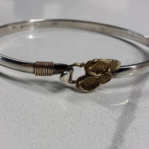 May include: Silver bangle bracelet with a gold-colored flip-flop design clasp. The bracelet has a polished finish and a hook closure. The clasp features two flip-flop shapes. The bracelet is a piece of jewelry.
