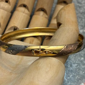 Peut inclure: Un bracelet jonc doré avec un fermoir décoratif. Le bracelet présente des sections alternées de tons or, argent et bronze avec des motifs de feuilles gravées. Le bracelet est présenté sur une main en bois.