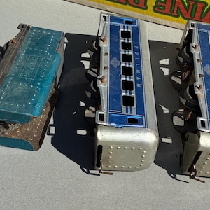 May include: A collection of vintage toy train cars in various states of wear. The cars feature a blue and red engine with a star design, and several silver and blue passenger cars. The train cars are made of metal and show signs of age.