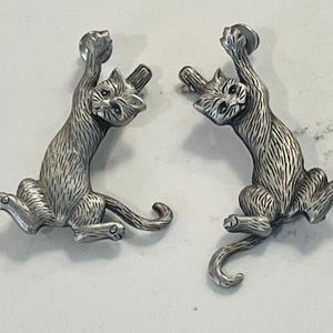 Op de afbeelding: Een paar zilveren oorringen in de vorm van een kat. De katten worden afgebeeld in een speelse pose, met hun poten in de lucht. De oorringen zijn een unieke en grillige toevoeging aan elke sieradencollectie.