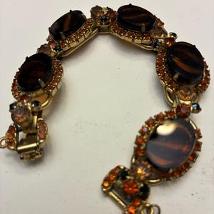 May include: Vintage gold-tone bracelet featuring oval-shaped, dark brown stones with a tiger-eye effect. The stones are surrounded by smaller orange and black rhinestones. A classic piece of jewelry.