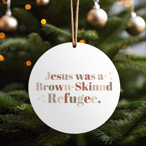 Può includere: Un ornamento natalizio rotondo e bianco con la scritta "Jesus was a Brown-Skinned Refugee" in tonalità marroni e grigie. L'ornamento è appeso a un cordino marrone sullo sfondo di un albero di Natale con palline e luci dorate.