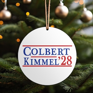 Peut inclure: Ornement de Noël rond et blanc avec l'inscription "COLBERT KIMMEL '28" en bleu et rouge. L'ornement est suspendu par une ficelle marron, sur un fond flou d'un sapin de Noël avec des boules et des lumières dorées.