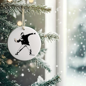 May include: A round, white Christmas ornament featuring a black silhouette of a person throwing an object. The ornament hangs from a beige string against a backdrop of a snowy window and a decorated Christmas tree.