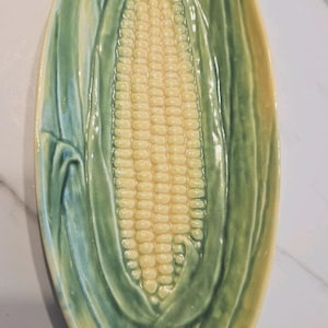 May include: Oval-shaped ceramic serving dish with a corn on the cob design. The dish features a yellow corn cob in the center, surrounded by green and yellow leaves. The dish is likely used for serving food.