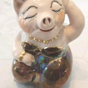 May include: A whimsical ceramic pig figurine with a light pink headpiece and a gold necklace. The pig is seated with its eyes closed and a serene expression. The lower half of the pig is iridescent brown. A unique decorative item.