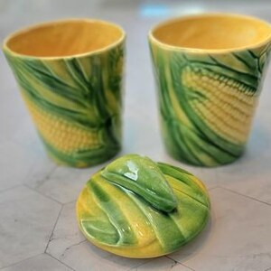 Puede incluir: Un juego de cerámica con temática de maíz que incluye dos tazas cilíndricas y una tapa. Las tazas tienen un interior amarillo y están decoradas con diseños de maíz verde y amarillo. La tapa es redonda con un asa en forma de hoja verde.