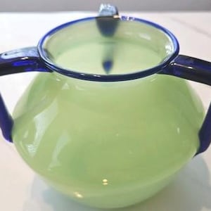 Puede incluir: Una jarra de vidrio verde claro con dos asas azul oscuro y un borde azul oscuro. La jarra tiene un cuerpo redondeado y un pequeño pico. El vidrio es liso y reflectante.