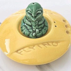Puede incluir: Un juego de salero y pimentero de cerámica. El juego presenta una base amarilla en forma de platillo con la palabra "FLYING" en relieve. Una figura de alienígena verde se encuentra en el centro. El juego tiene una estética retro y kitsch.