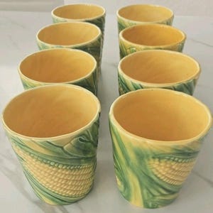 May include: A set of eight ceramic tumblers. The tumblers have a yellow interior and are decorated with a corn-on-the-cob design in shades of yellow and green. The exterior features embossed corn kernels and leaves.