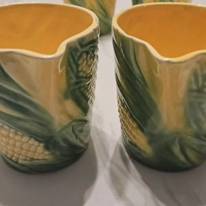 May include: Two ceramic corn-shaped vases. The vases have a yellow interior and are decorated with green leaves and corn kernels. The design is a detailed, three-dimensional relief. The vases are likely for decorative purposes.