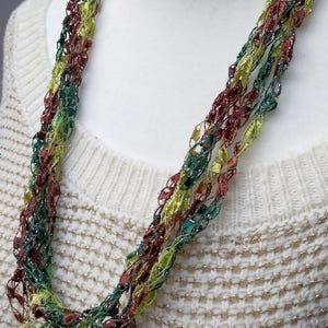 Puede incluir: Un collar de múltiples hebras con un diseño tejido. El collar presenta hebras de hilos verdes, amarillos y granates. El collar se muestra en un maniquí blanco. La modelo lleva un suéter color crema.