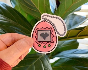 May include: A pink and gray Tamagotchi keychain with a heart on the screen. The keychain is made of vinyl and has a silver chain.