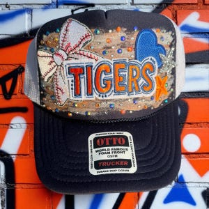 Może przedstawiać: Czarna i biała czapka typu trucker z napisem "TIGERS" w pomarańczowych i białych literach. Czapka posiada wzór baseballu i kokardy, niebieskie serce i gwiazdę. Czapka ma piankowy przód i zapięcie na zatrzask.