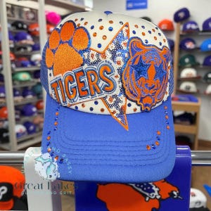 Könnte beinhalten: Eine blau-weiße Baseballkappe mit dem Wort "TIGERS" in orangefarbenen Buchstaben. Die Kappe hat einen orangefarbenen Pfotenabdruck, einen Stern und ein Tigerkopf-Design. Der Schirm ist blau und die Kappe ist mit Strasssteinen verziert.
