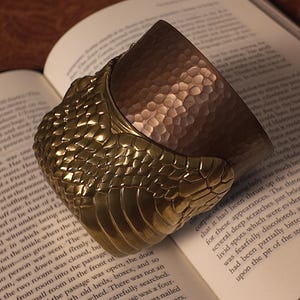 May include: A unique book weight featuring a detailed, textured design. The top portion has a hammered copper finish, while the base is a gold-toned, scale-like pattern. It rests on the open pages of a book.