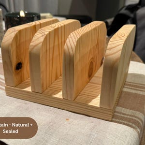 Puede incluir: Un servilletero de madera natural de color claro con cuatro divisores verticales con bordes superiores redondeados. La veta de la madera es visible en toda la estructura, sellada sin tinte. El texto en la imagen dice: No Stain - Natural + Sealed.