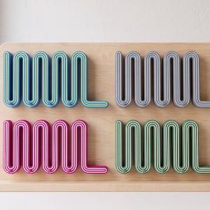 Op de afbeelding: Een lichtgekleurd houten displaybord met vier afzonderlijke wandhaken, elk gevormd als een doorlopend, herhalend golf- of zigzagpatroon. De haken zijn gekleurd in blauw, grijs, magenta en groen, elk met contrasterende binnenlijnen.