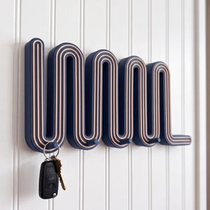 May include: A wall-mounted coat rack featuring a continuous, wavy, serpentine design in dark navy blue with light beige layered edges. The rack includes four small pegs for hanging items, with a black car key fob hanging from the first peg.