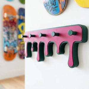 May include: A wall-mounted coat rack featuring a drip-effect design in magenta and dark green. The horizontal base has five cylindrical pegs extending outward, with the bottom edge shaped like irregular liquid drips in a solid colour scheme.