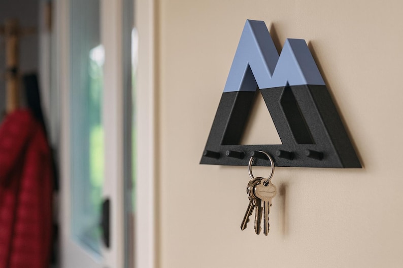 Black Mountain key holder with blue-gray peaks holding house keys on a beige wall, a red jacket by the door behind