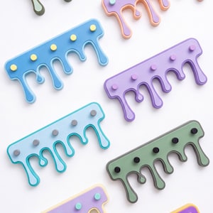 May include: Eight wall-mounted coat racks featuring a dripping paint design. Each rack has a rectangular base with four circular pegs and four downward-extending drips in various colour combinations, including sage green, lavender, blue, and yellow.