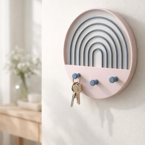May include: A round, pink key holder mounted on a white wall. The key holder features a blue rainbow design and three blue pegs for hanging keys. Two keys are hanging from one of the pegs. A vase of flowers is visible in the background.
