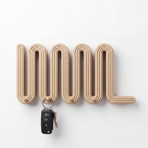 May include: A light beige wall-mounted coat rack featuring a continuous, repeating serpentine wave pattern with integrated hooks. The rack is constructed with multiple parallel ridges and holds a black electronic car key fob on one of the hooks.