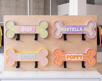 Può includere: Quattro ganci da parete a forma di osso montati su una tavola di legno chiaro. Ogni gancio presenta linee ondulate colorate e stratificate, con una placca rettangolare centrale che riporta i nomi GIGI, STELLA, LUNA e POPPY in rilievo.
