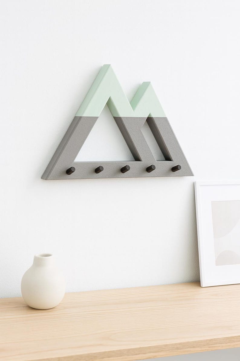 Gray Mountain key holder with mint green peaks on a white wall above a light wood shelf, a white bud vase and leaning frame