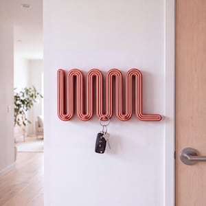 May include: A terracotta-colored wall-mounted key holder featuring a continuous, repeating wavy line design with integrated hooks. A silver metal keyring with a black car key fob hangs from one of the hooks on the textured, ribbed surface.
