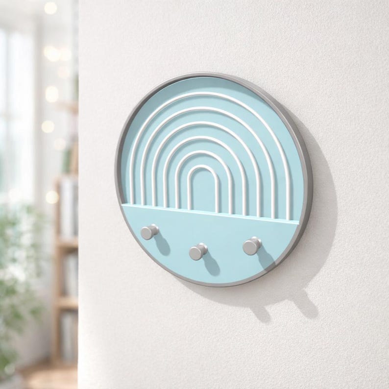 Pale blue Boho key holder casting a soft shadow on a white wall, pegs empty, a sunny window and bookshelf blurred at left