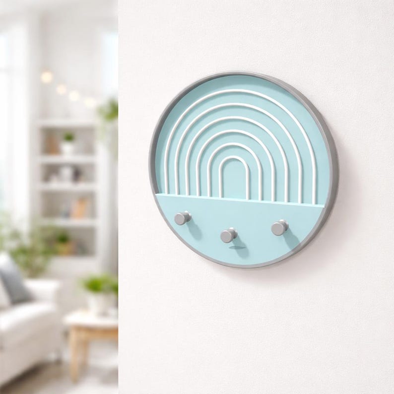 Pale blue Boho key holder angled on a white wall, three gray pegs empty, a bright living room with shelves and sofa behind