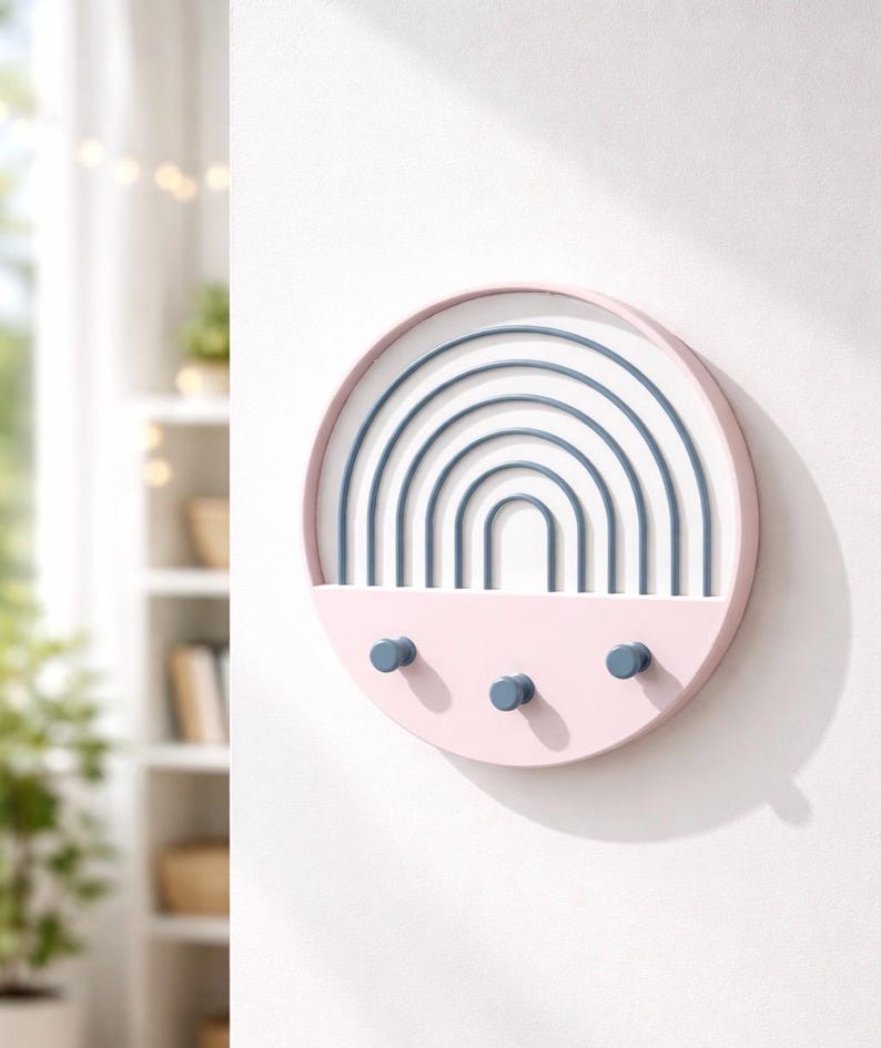 Pink Boho key holder with blue rainbow arches on a sunlit white wall, a bookshelf with plants and string lights behind