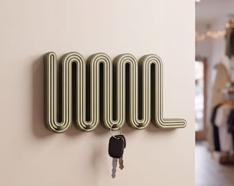 May include: A wall-mounted olive green coat rack featuring a continuous, repeating serpentine wave pattern with integrated hooks. The structure has a layered, ribbed texture and holds a small metal ring with two silver-colored keys.