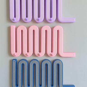 Puede incluir: Tres percheros de pared ondulados en colores pastel: lavanda, rosa y azul. Cada gancho tiene un diseño único con una serie de líneas curvas. Los ganchos están montados en una pared gris claro, ofreciendo una solución de almacenamiento moderna y decorativa.