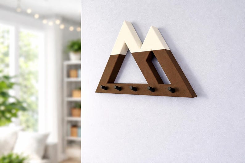Brown and white Mountain key holder on a white wall, five black pegs empty, a bright room with shelves blurred at left