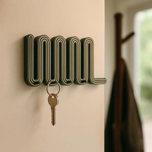 May include: A dark green key holder mounted on a white wall. The key holder has a unique, wavy design with white accents. A set of keys hangs from the holder, suggesting its practical use for organizing keys. The design is modern and minimalist.