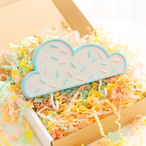May include: A cloud-shaped wall hook with a light blue border and white center, decorated with pink and blue sprinkles. The hook has three white hooks and is presented in a cardboard box filled with colorful paper shreds.