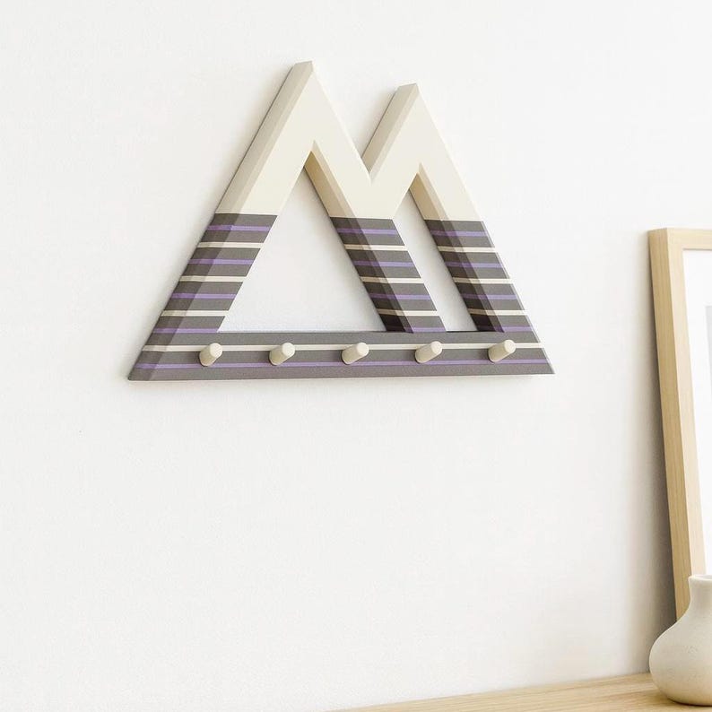 Multi-colored Mountain key holder with cream peaks and gray and purple stripes on a white wall, a wood frame and vase nearby