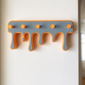 May include: A wall-mounted coat rack with a unique design. The rack is gray with orange accents and has five orange pegs. The bottom edge has a dripping paint effect. The rack is mounted on a white wall.