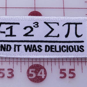 May include: A white rectangular patch with black embroidered text and symbols. The text includes mathematical symbols and the phrase "AND IT WAS DELICIOUS". The patch is on a grid background with inch measurements.