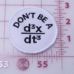 Pode incluir: Um patch circular branco com texto bordado em preto. O texto diz "DON'T BE A" acima de uma equação matemática, "d³x/dt³". O patch tem uma borda costurada e está sobre um fundo quadriculado.