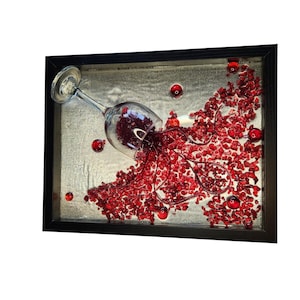 Pode incluir: Uma obra de arte emoldurada retrata um copo de vinho derramando líquido vermelho e pequenas contas vermelhas. O vidro é transparente e o fundo é de cor prateada texturizada. Várias contas vermelhas estão espalhadas.